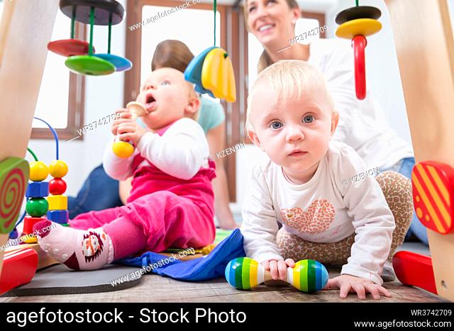 Cute baby girl showing progress and curiosity by trying to reach multicolored wooden toys, while sitting on the floor with her mother and a family friend
