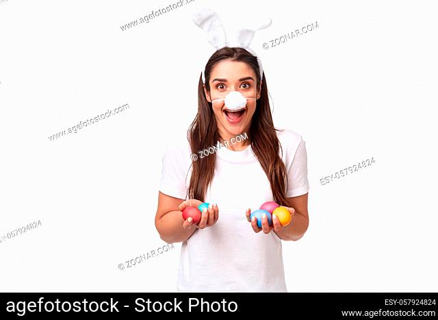 Portrait of excited, happy and carefree young woman spending holiday with family, wearing funny rabbit ears and nose, holding colored eggs