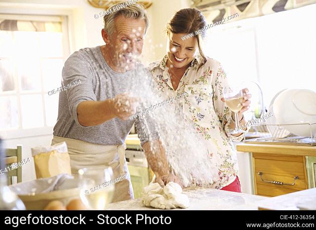 Playful mature couple baking, throwing flour and drinking wine in kitchen