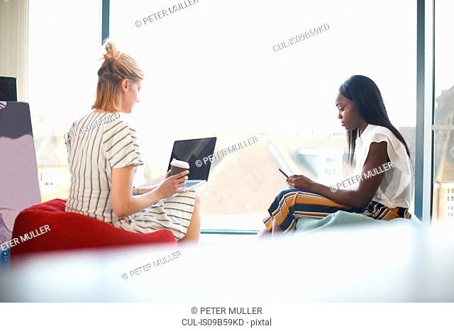 Two businesswomen sitting on beanbags looking at laptop and smartphone