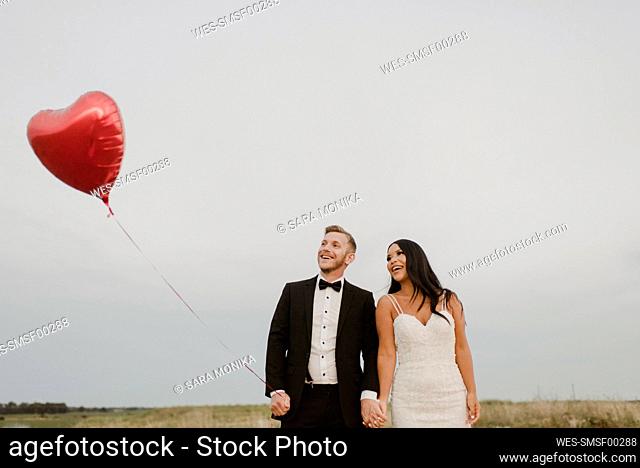 Happy bride and groom with heart shape balloon against sky