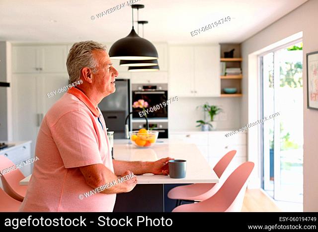 Relaxed caucasian senior man sitting in kitchen with coffee and looking through window