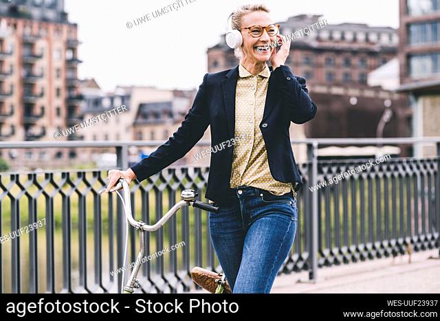 Happy female professional listening music through headphones while wheeling bicycle on bridge