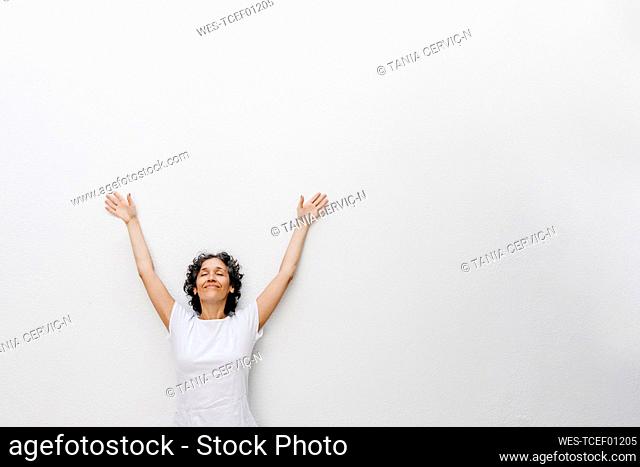 Smiling mature woman standing with eyes closed and arms raised against white wall