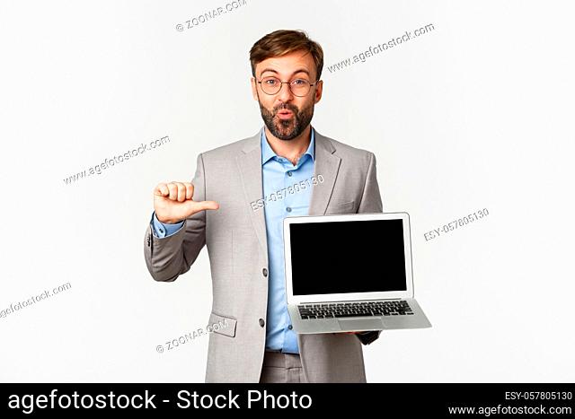 Portrait of happy bearded businessman in glasses and gray suit, pointing at laptop screen and looking amazed, demonstrating something good