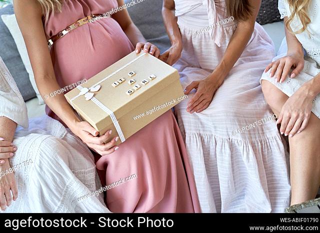 Hands of pregnant woman opening gift box sitting with friends at home