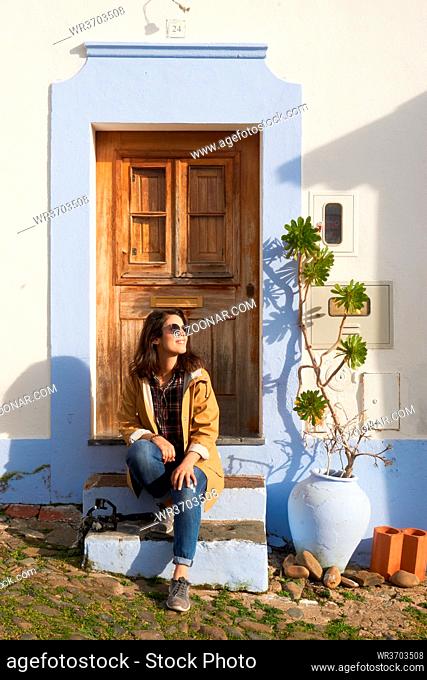 Woman girl tourist traveler with yellow jacket sitted on the stairs of a cute picturesque antique white and blue house with a plant in Mertola Alentejo