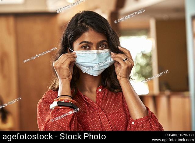 Portrait of young ethnic woman putting on face mask wearing orange blouse sitting at bar in kitchen of downtown loft