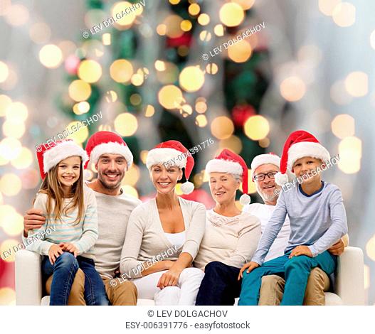 family, happiness, generation, holidays and people concept - happy family in santa helper hats sitting on couch over christmas tree lights background