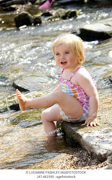 18 month old baby girl, sitting on a stone in a river kicking her foot in the air, smiling