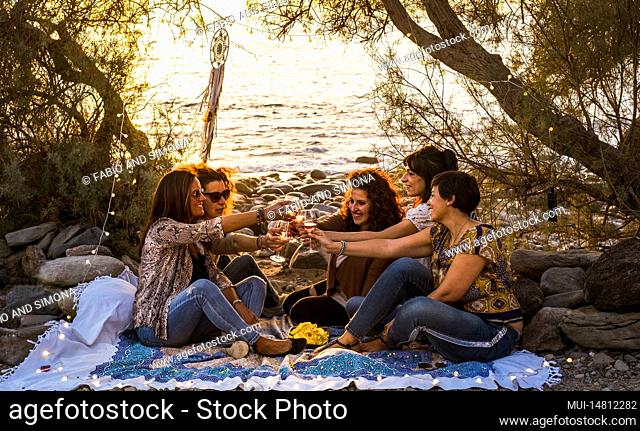 Group of females young middle age friends celebrate all together sitting at the natural rocks beach and drinking wine toasting and smiling