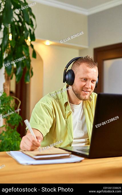 Happy Man wearing wireless headphones watch webinar, trainning, course, lessons on laptop take notes to paper notebook at home