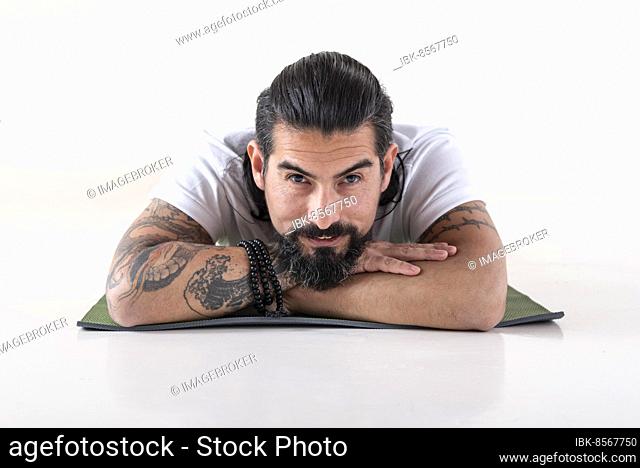 Portrait of a man dressed in white yoga clothes lying on a mat while looking at camera over white background. Studio shot