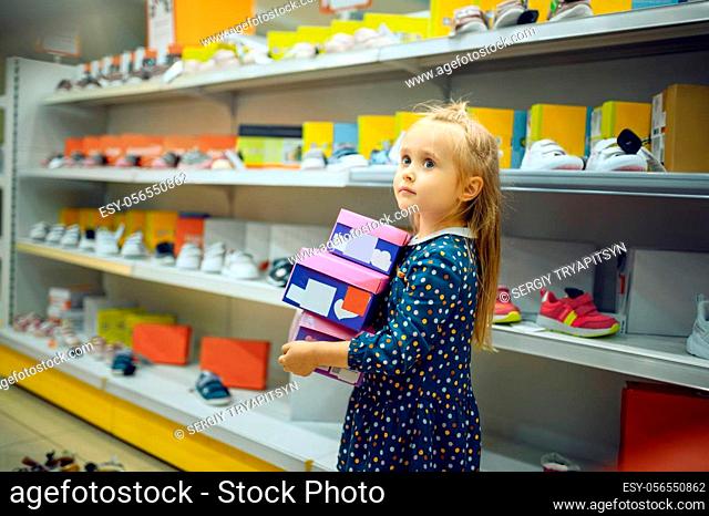 Pretty little girl holds box with shoes in kid's store. Adorable child looking on showcase in children's shop, happy childhood, baby makes a purchase in market