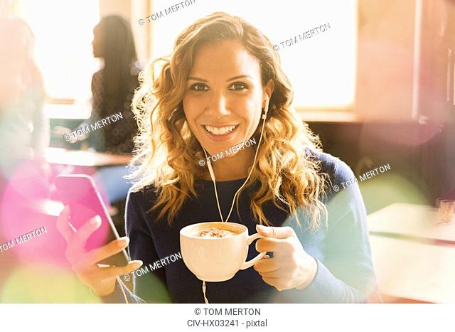 Portrait smiling woman with headphones listening to music on mp3 player and drinking cappuccino in cafe