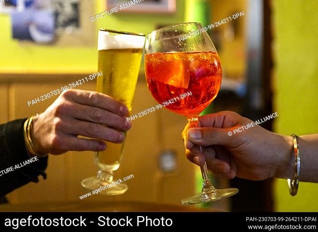PRODUCTION - 01 July 2023, Berlin: In a bar, two visitors toast with a glass of non-alcoholic beer and a glass of Aperol Spritz