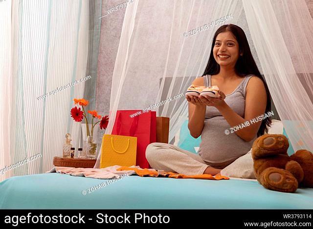 Smiling pregnant woman holding a pair of baby shoes while sitting on bed at home