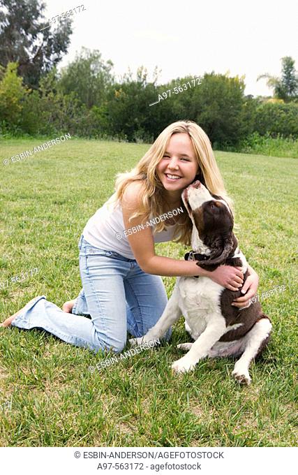 Smiling blonde teen girl in blue jeans and white tank top kneeling on grass lawn, get's a kiss from her pet dog
