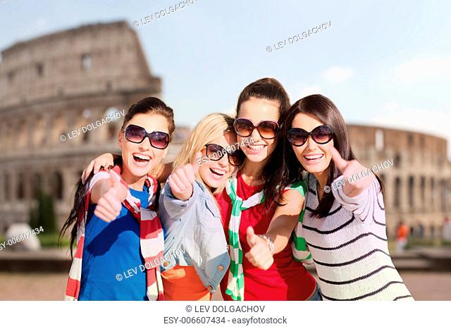 summer holidays, vacation, travel, friendship and people concept - happy teenage girls or young women in sunglasses showing thumbs up and laughing over coliseum...