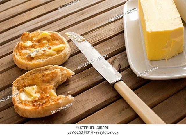 One yellow stick of butter on a white tray beside a knife and two toasted english muffins set on table made of wooden slats