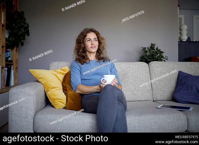 Smiling with coffee cup woman looking away while sitting on sofa at home