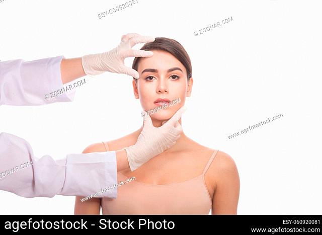 Handmade beauty. Portrait of beautiful young woman keeping eyes opened while professional doctor in medical gloves touching her face isolated on white...