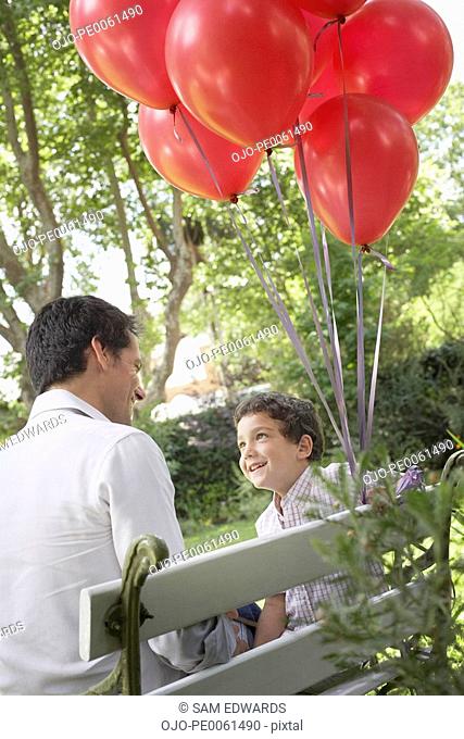 Man outdoors sitting on bench with young boy beside him holding red balloons smiling