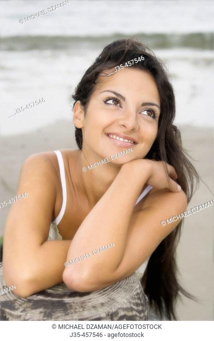 A smiling young girl 20's to 30's with long dark hair, leans her arms on a pier post during a breezy summers day