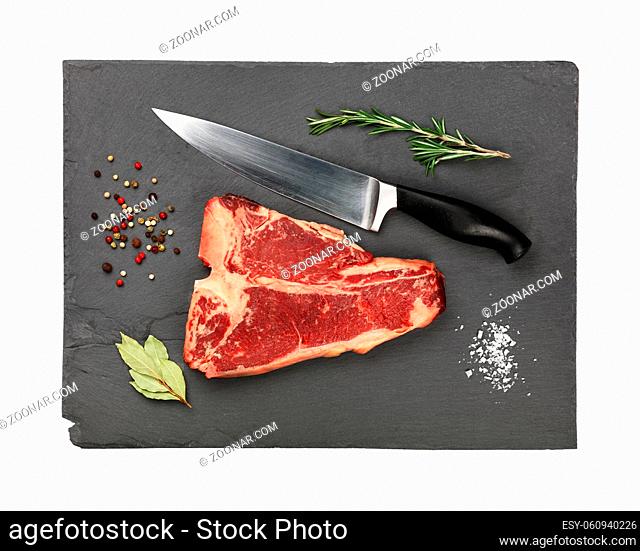 Close up one marbled raw porterhouse T-bone beef steak with rib bone on black slate cutting board with knife and spices to season, isolated on white background