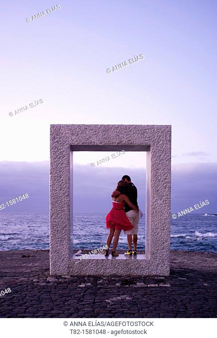 Young couple embracing in a square geometric shape sculpture by the sea, Garachico, Tenerife North, Canary Islands, Spain