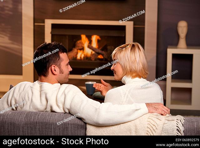 Young romantic couple sitting on sofa in front of fireplace at home, looking at each other, talking, rear view