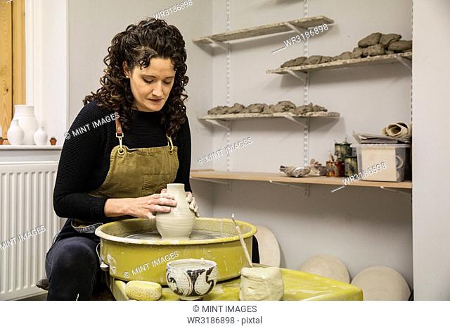 Woman with brown curly hair wearing apron shaping clay vase on pottery wheel