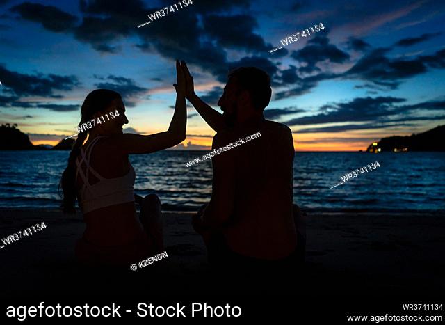 Cheerful young couple giving high five while sitting on a tropical beach at dusk during summer vacation in Flores Island, Indonesia