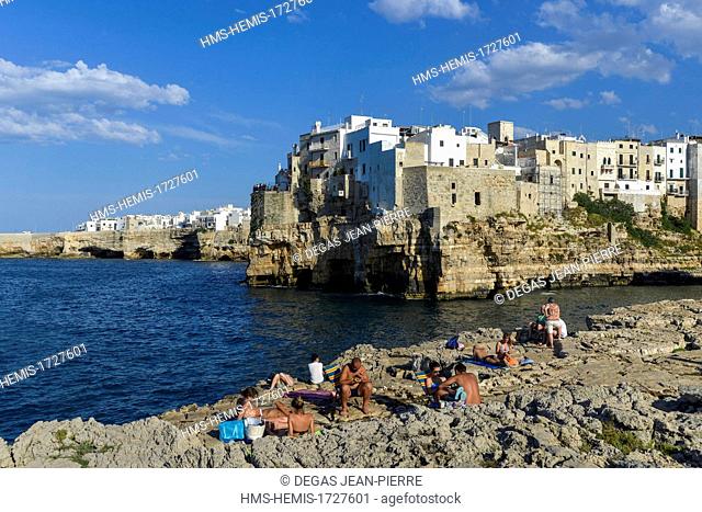 Italy, Puglia, Bari province, Polignano a Mare, vacationers on deckchairs taking the sun in front of the old town built on an abrupt cliff which dominates a...