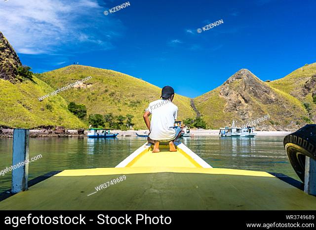 Rear view of a young man enjoying the tranquil view of Padar Island, while sitting on a jetty in a sunny day during summer vacation in Indonesia
