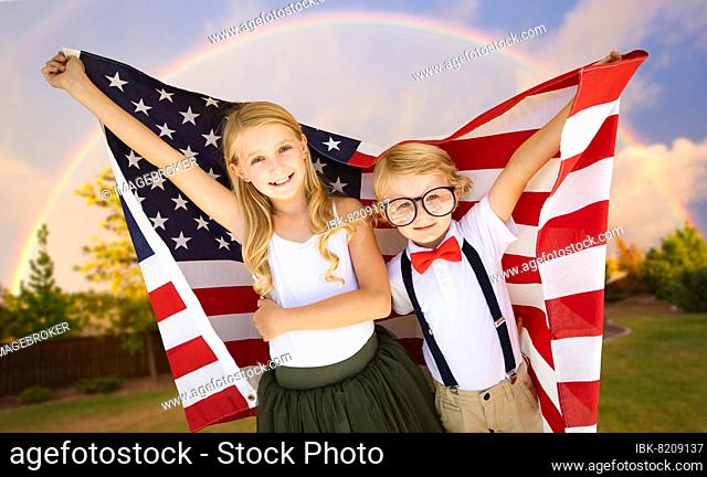 Cute young cuacasian boy and girl holding american flag with rainbow behind