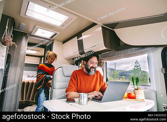 Daily normal life inside a camper van rv mobile home. Woman cooking in the kitchen and adult man working on laptop computer