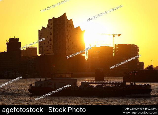07 October 2022, Hamburg: The sun rises atmospherically between construction cranes in the morning behind the Elbphilharmonie concert hall in Hamburg harbor