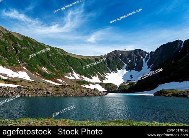 Stunning summer mountain landscape of Kamchatka Peninsula: Blue Lake, snow and ice along shores of mountain lake, green forest on hillsides