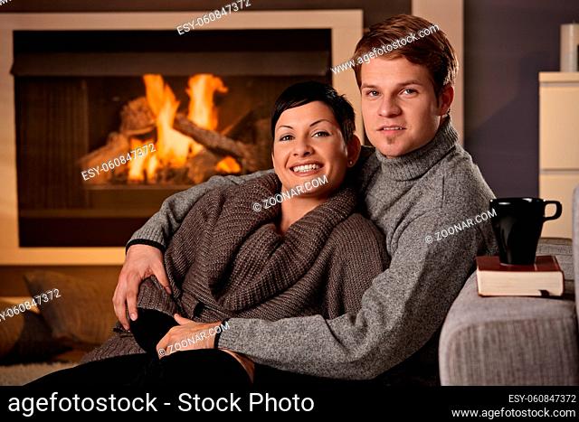 Young couple sitting on floor hugging in front of fireplace at home, looking at camera, smiling