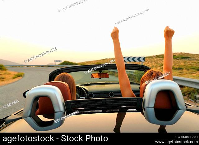 Back view of two excited women driving a convertible car in the road on summer vacation