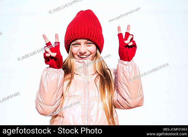 Smiling girl in warm clothing showing Peace Sign against wall