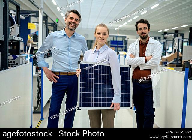 Smiling young engineer holding solar panel standing with colleagues at factory