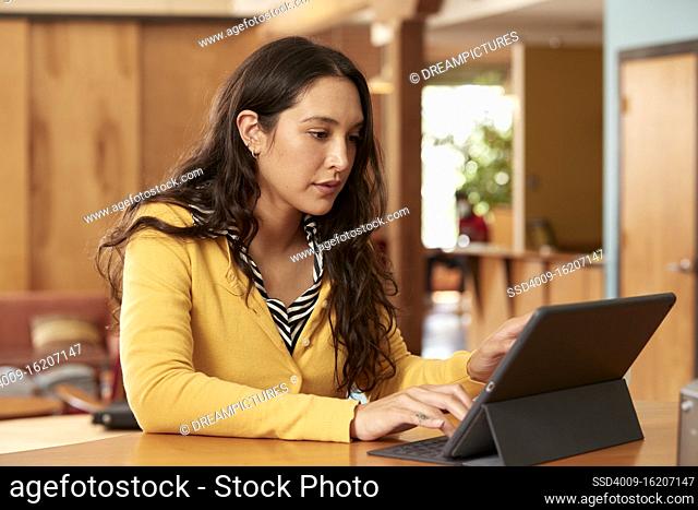 Portrait of young ethnic woman wearing yellow sweater with black and white striped blouse, sitting at bar in kitchen of downtown loft with iPad