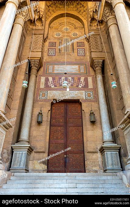 Side entrance of al Rifai Mosque, old decorated bricks stone wall with arabesque decorated wooden door framed by marble engraved cylindrical columns