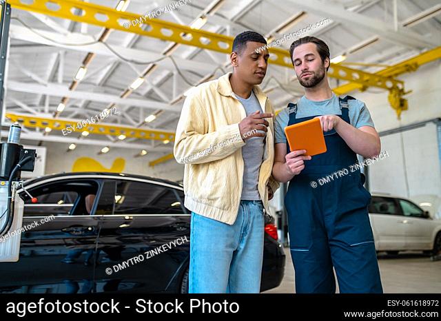 Good offer. Man in work overalls showing tablet screen to dark skinned customer standing chatting in auto repair shop