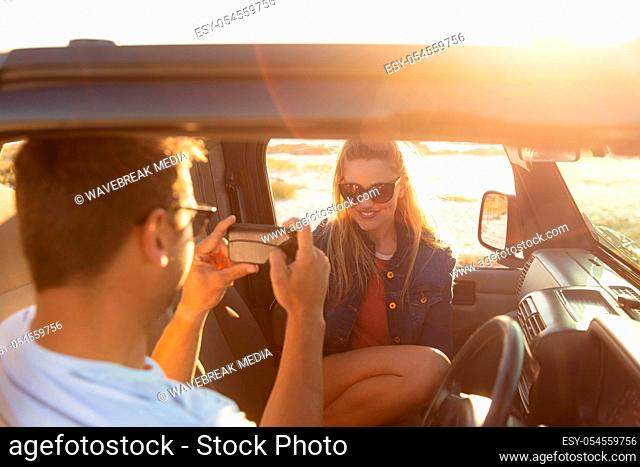 Rear view of a Caucasian man with his girlfriend smiling inside an open top car, taking a photo of her