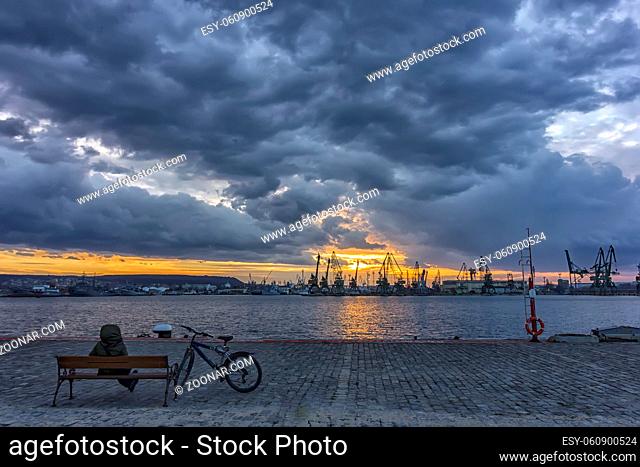 a man with a bike sitting on a bench in a harbor to admire the sunset/sunrise