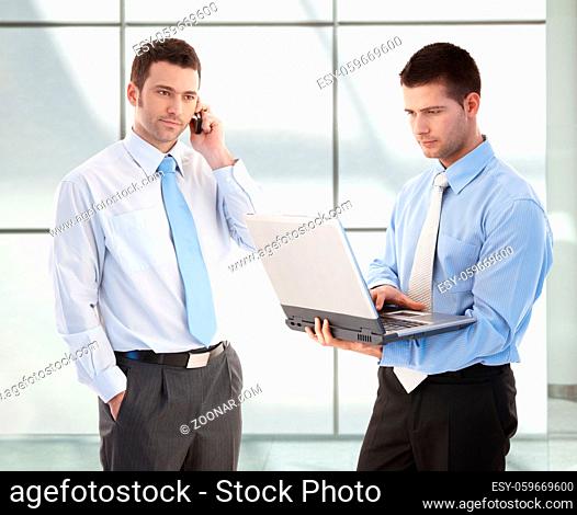 Young businessmen standing in office lobby, one on phone, the other working on laptop