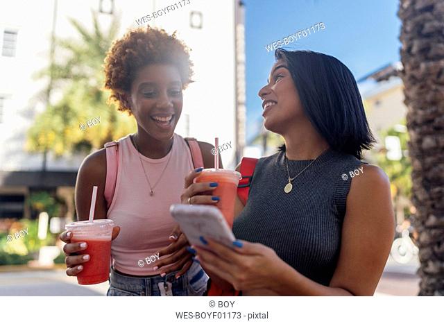 USA, Florida, Miami Beach, two happy female friends with cell phone and soft drink in the city
